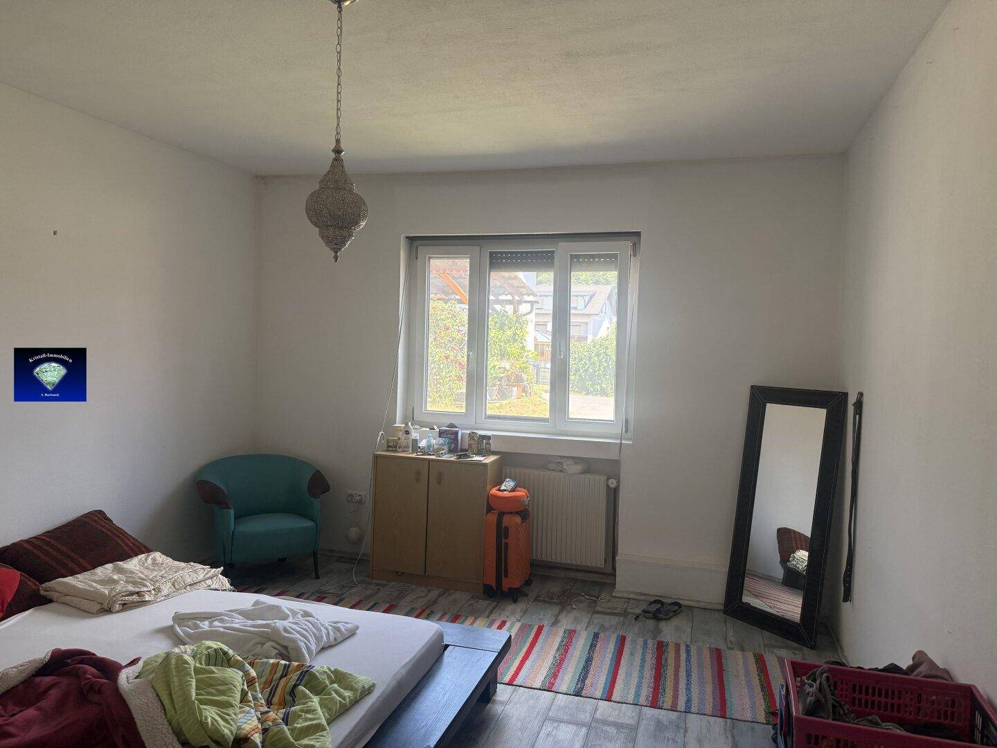 Schlafzimmer mit einem Bett, einem Sessel und einem großen Fenster, das viel Tageslicht hereinlässt.