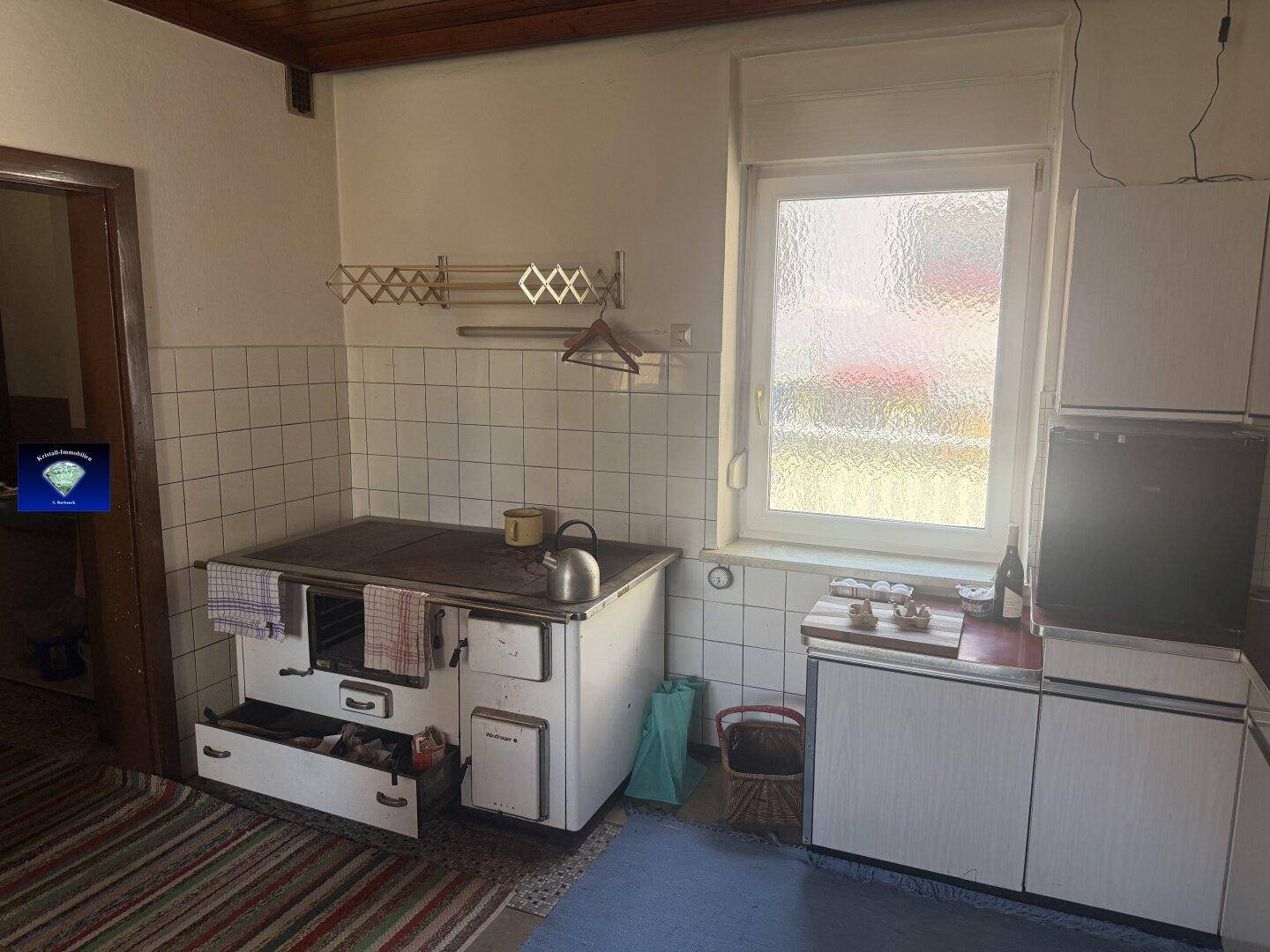 Einfache Küche mit einem traditionellen Holzofen, weißen Fliesen und einem Fenster mit Milchglas.
