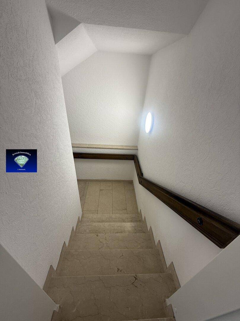 Marmortreppe mit Holzgeländer führt nach unten, gut beleuchtet durch Deckenlicht.