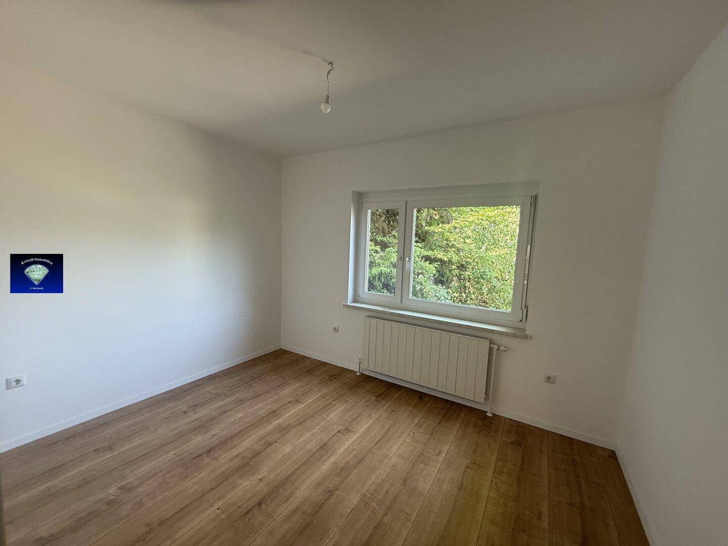 Helles Zimmer mit Laminatboden und Fenster, das einen grünen Ausblick bietet.