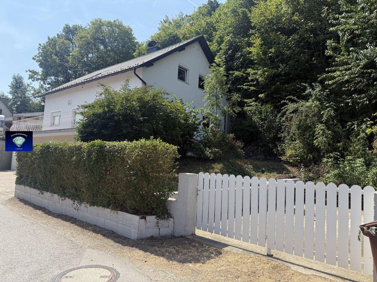 Seitenansicht des Hauses mit weißem Lattenzaun und Hecke, umgeben von Bäumen.