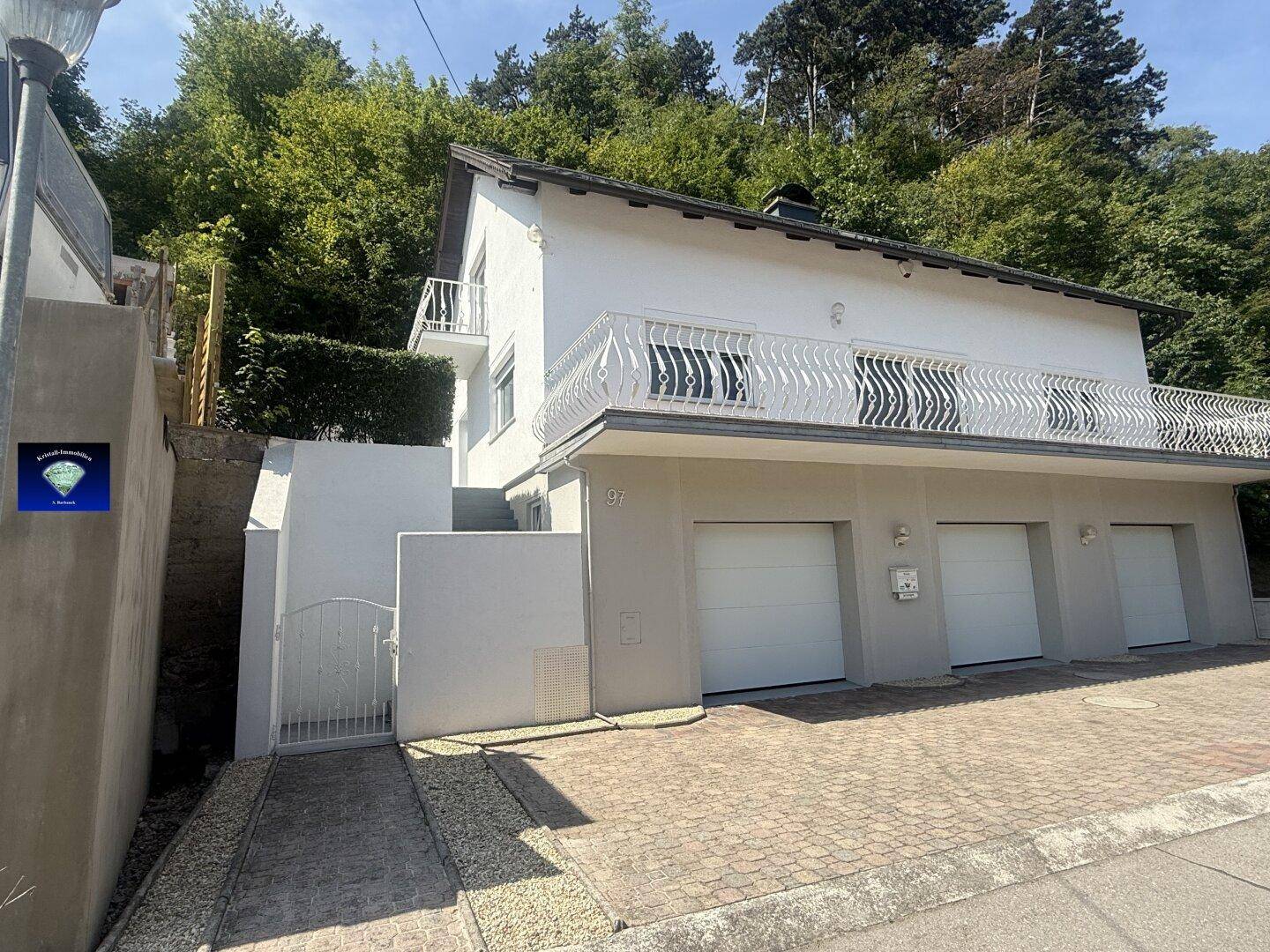 Ansicht des Hauses mit Garagen und Balkon, eingebettet in eine grüne Hanglage.