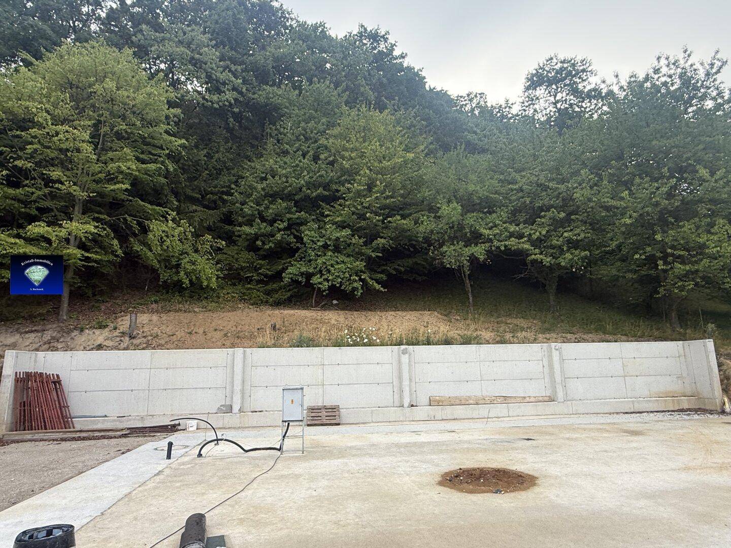 Baustelle mit langer Betonstützmauer, vorbereiteter Bodenplatte und Wald am Hang.