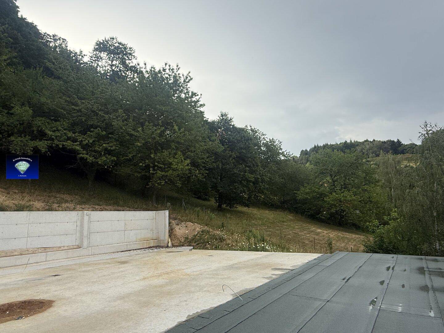 Baustelle mit Betonbodenplatte, teilweise gedecktem Dach und bewaldetem Hang im Hintergrund.
