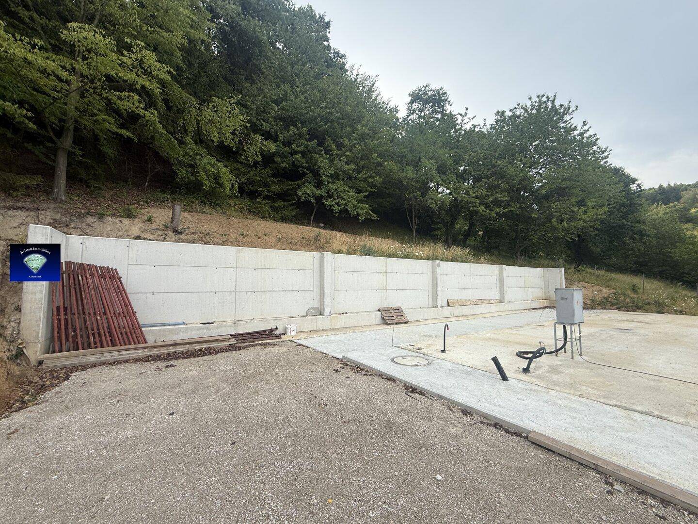 Baustelle mit einer langen Betonstützmauer und einer großen Betonfläche vor bewaldetem Hang.