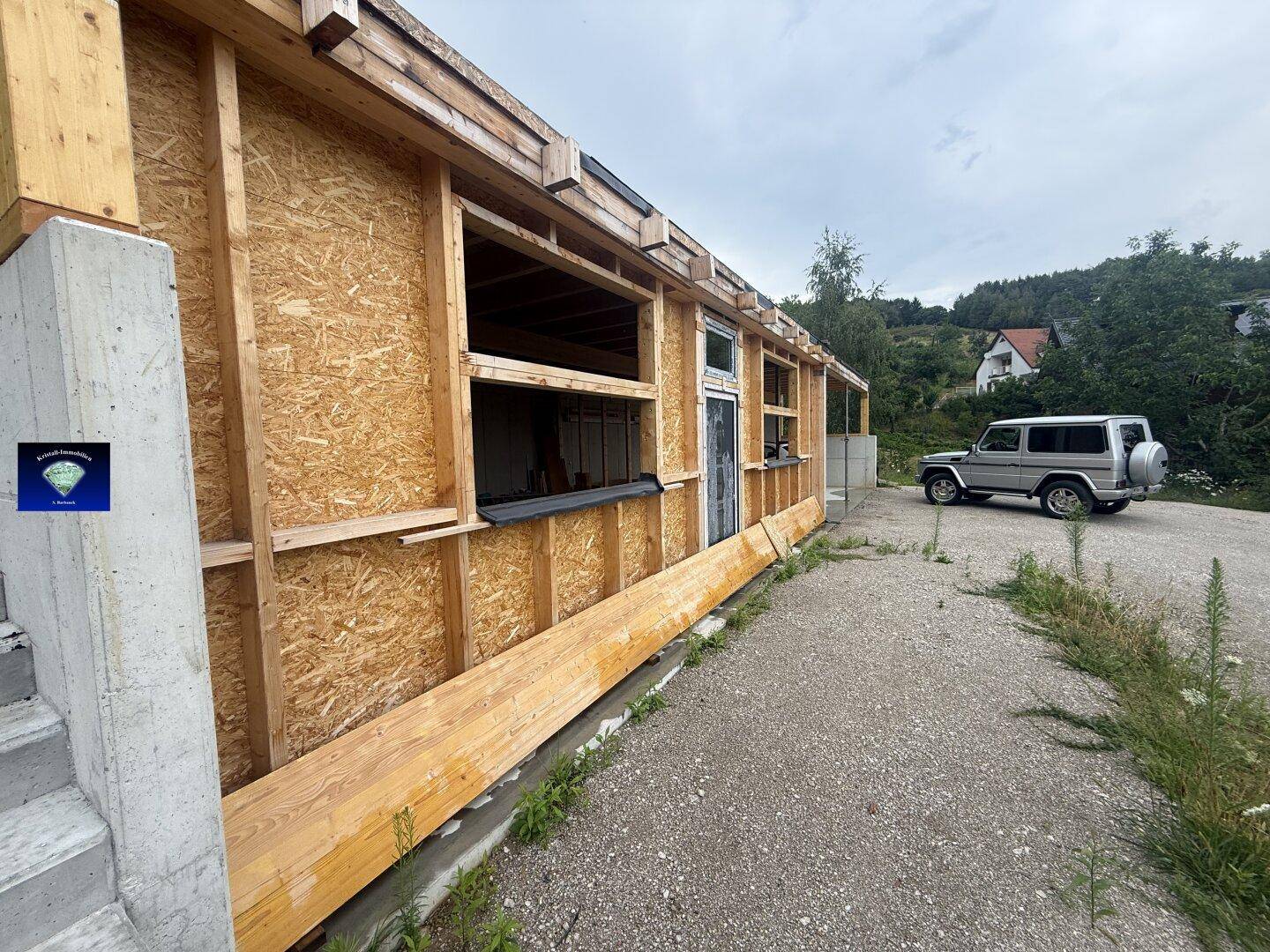 Außenansicht eines Hauses im Bau mit Holzrahmenwänden und Fensteröffnungen, ein Auto parkt daneben.