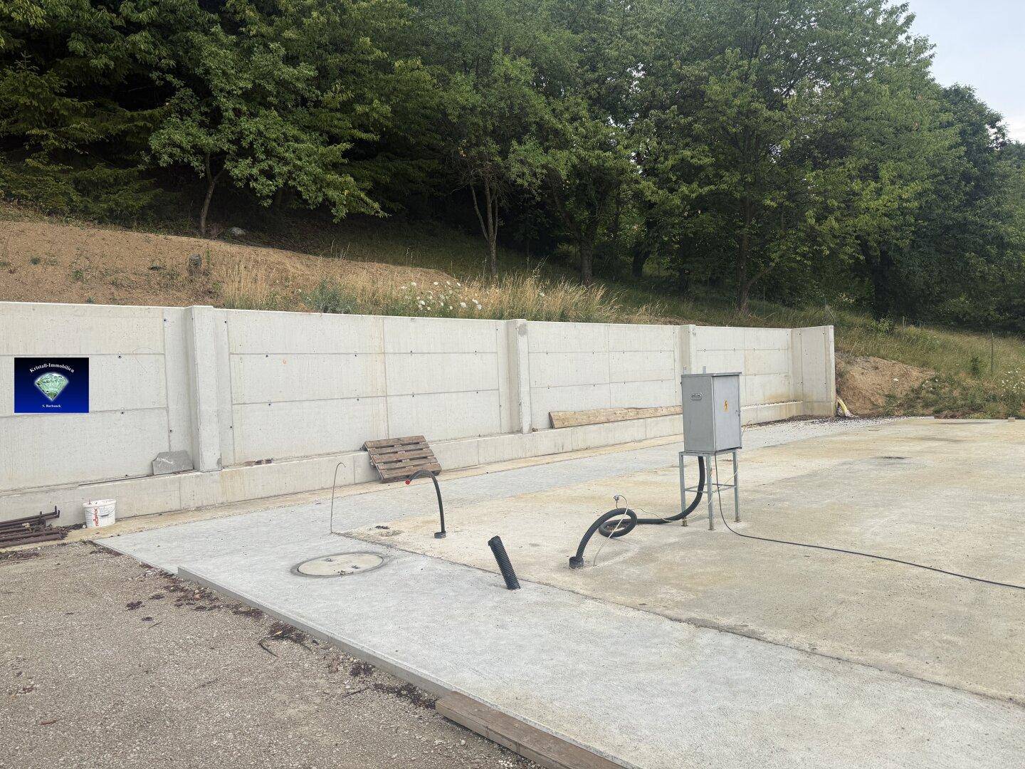 Baustelle mit Betonstützmauer, vorbereiteter Bodenplatte und sichtbaren Installationen am Hang.