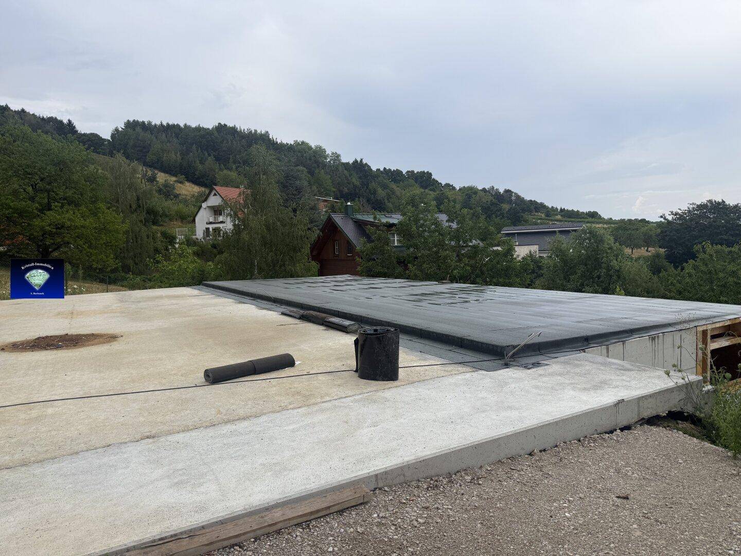 Baustelle mit Betonbodenplatte und teilweise gedecktem Flachdach, Häuser in der Ferne sichtbar.