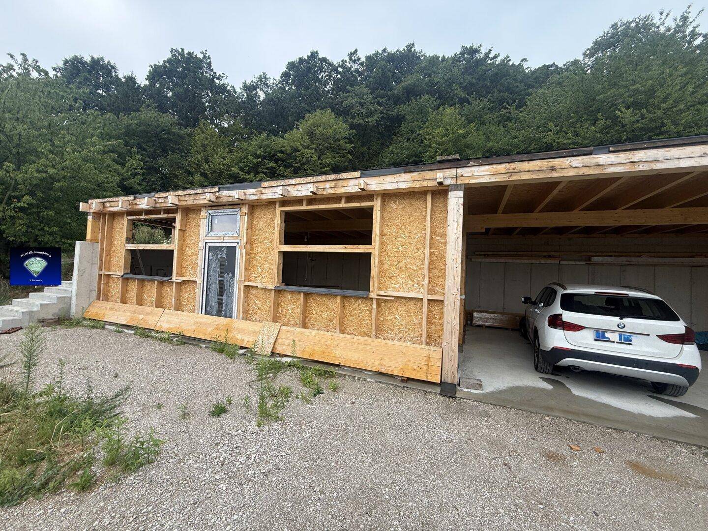 Außenansicht eines Hauses im Bau mit Holzrahmenwänden, Fensteröffnungen und einem geparkten Auto.