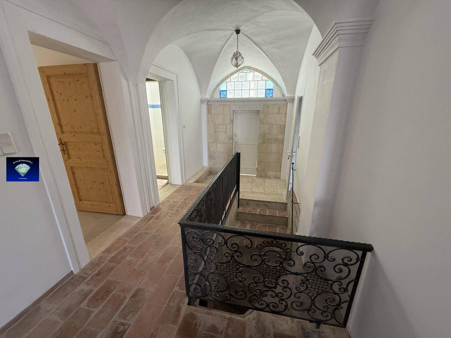 Historischer Flur mit Terrakottafliesen, Gewölbedecke und kunstvollem schmiedeeisernem Geländer, führt zu Treppen.