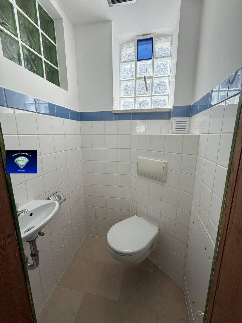Kleines WC mit weißen Fliesen, wandhängender Toilette, kleinem Waschbecken und zwei Fenstern.
