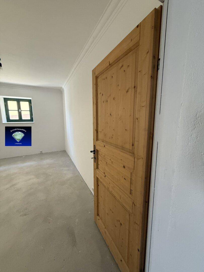 Rustikale Holztür in einem leeren Raum mit Betonboden und kleinem Fenster, schlicht und funktional.