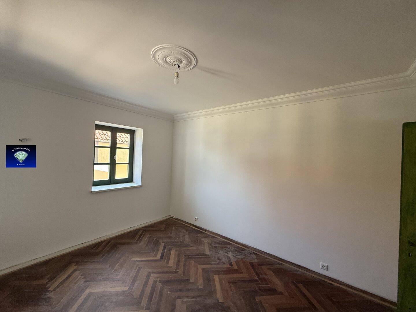 Heller Raum mit Fischgrätparkettboden, weißen Wänden und einem grünen Fenster, inklusive Deckenrosette.