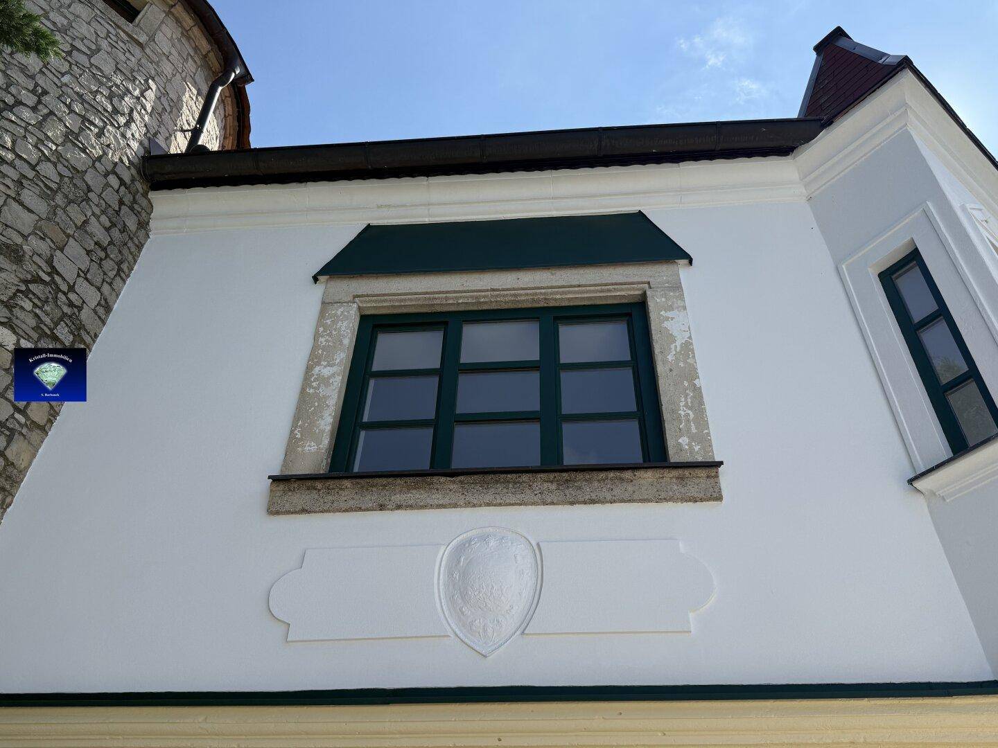 Weiße Fassade mit einem grünen Fenster und dekorativen Stuckelementen unter einem dunklen Vordach.