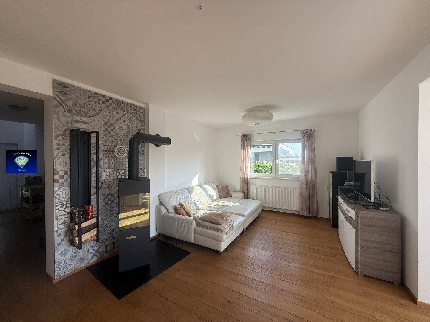 Gemütliches Wohnzimmer mit Holzboden, Kaminofen und bequemer Couch, hell beleuchtet.
