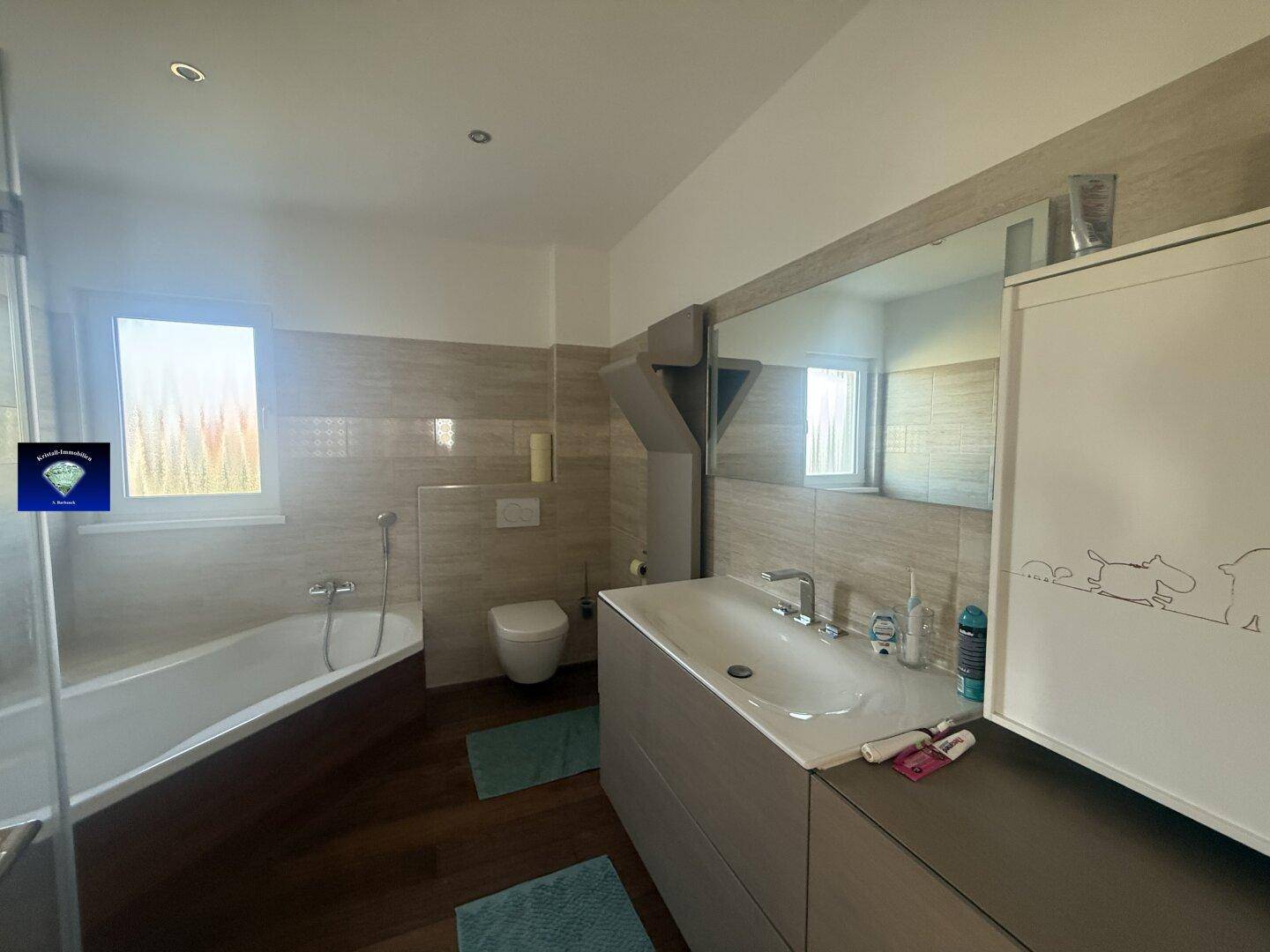 Helles Badezimmer mit großem Spiegel, Waschtisch und Badewanne unter dem Fenster.