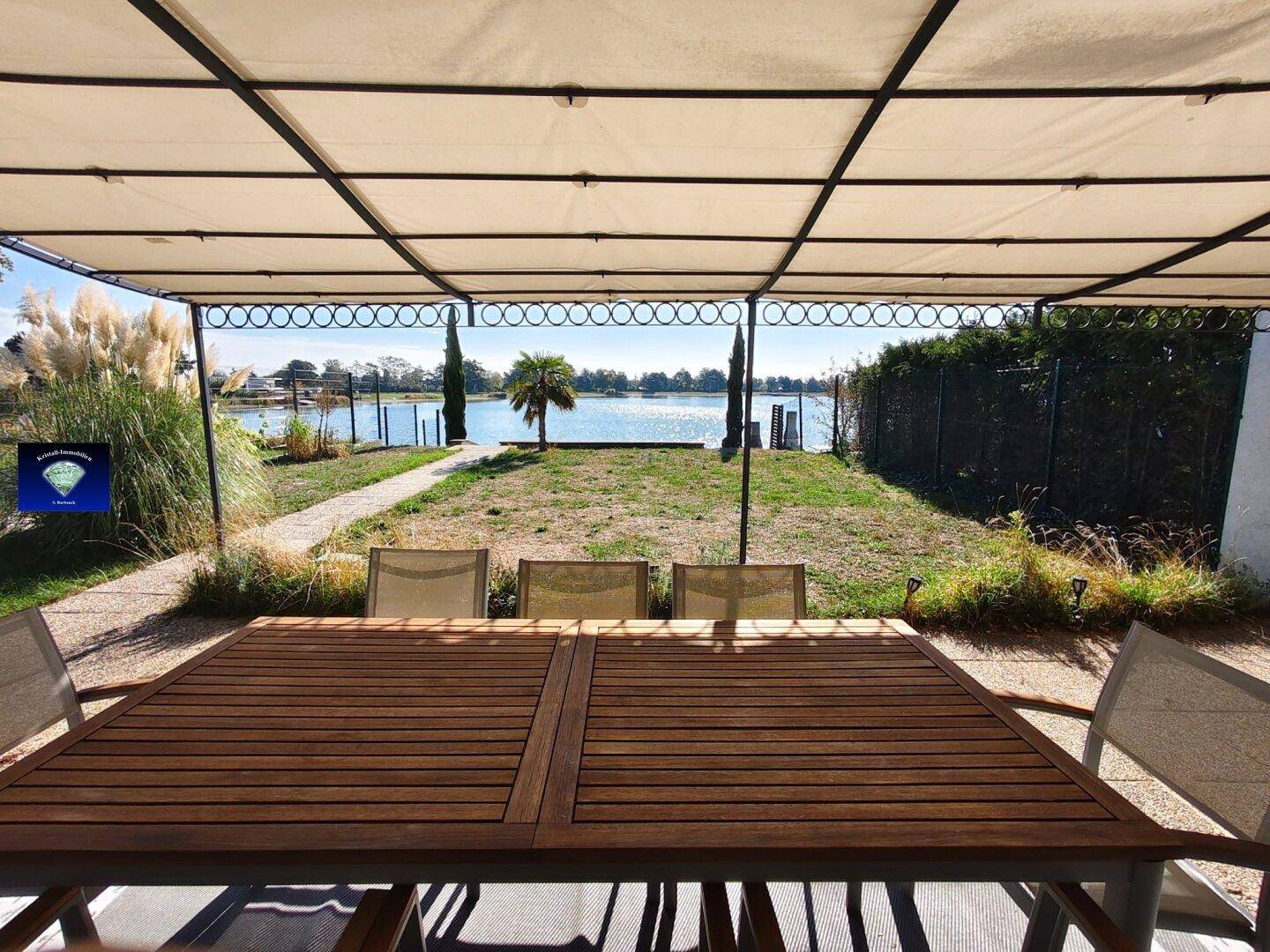 Blick von der überdachten Terrasse auf den See, mit Esstisch im Vordergrund und Palmen am Ufer.