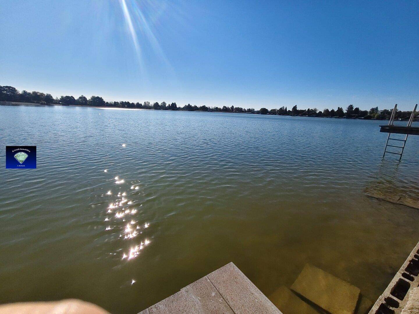 Weiter Blick über den sonnigen See mit einem kleinen Steg und Leiter am Ufer.