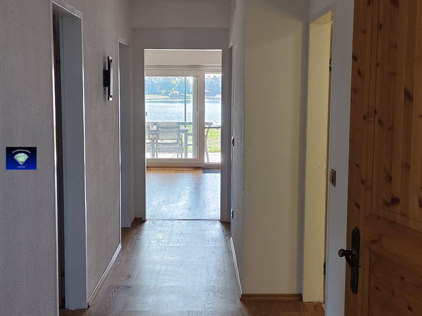 Heller Flur mit Holzboden, der zum Wohnbereich mit Seeblick durch die Glastüren führt.