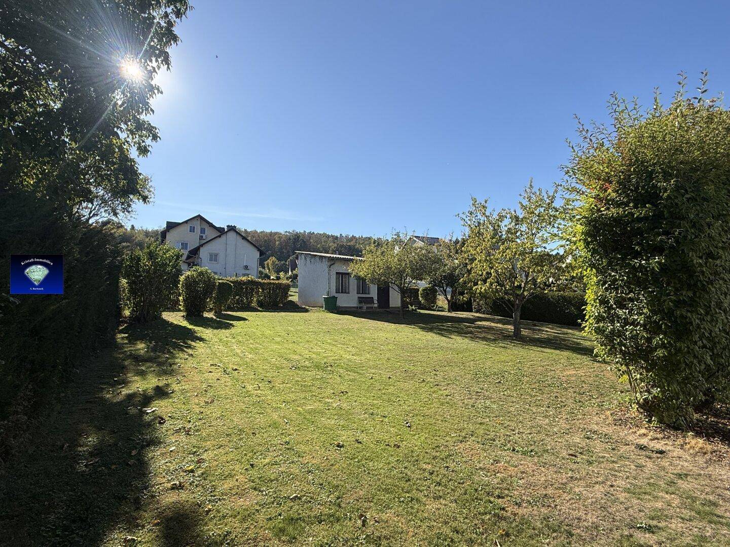 Weitläufiger Garten mit Rasen, Bäumen und einem kleinen Nebengebäude im Hintergrund bei Sonnenschein.