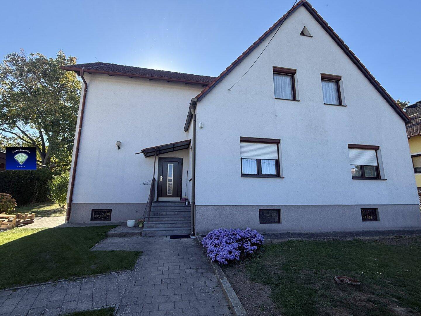 Vorderansicht eines weißen Einfamilienhauses mit braunem Dach, Fenstern mit Rollläden und Vorgarten.
