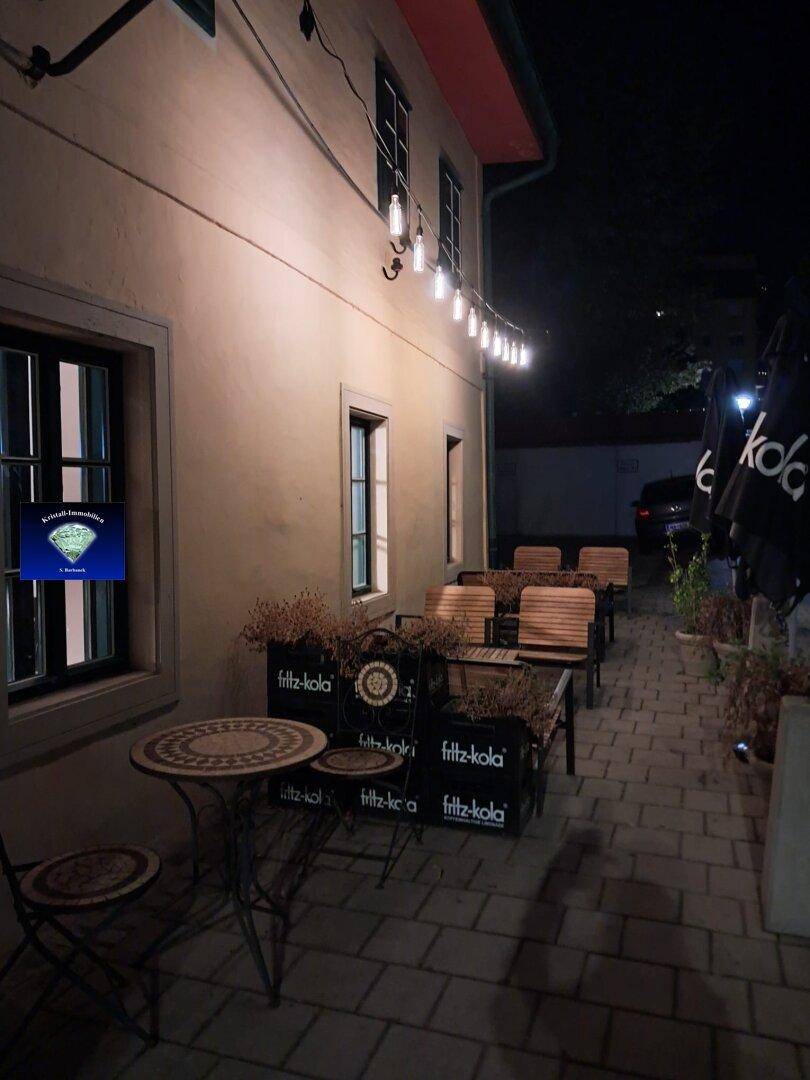 Gemütliche Außenterrasse mit Holztischen, Bänken und stimmungsvoller Lichterkette am Abend.