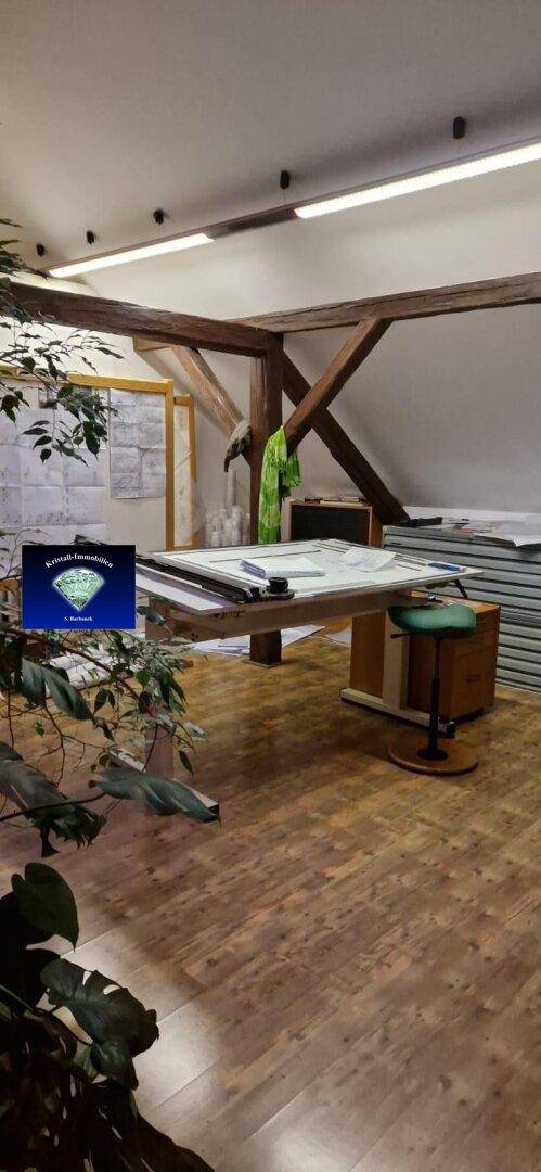 Helles Dachgeschossbüro mit sichtbaren Holzbalken, Holzboden und einem großen Zeichentisch.