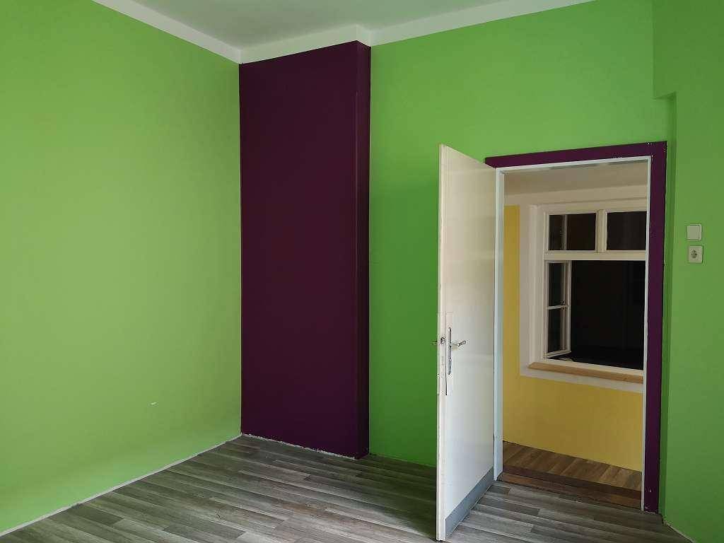 Zimmer mit grünen und violetten Wänden, einem Holzboden und einer offenen Tür zu einem weiteren Raum.