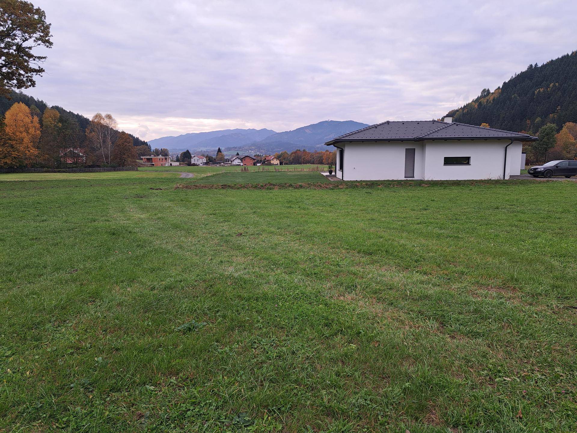 Modernes weißes Haus auf einer weitläufigen Wiese mit Blick auf ein Dorf und Berge in der Ferne.