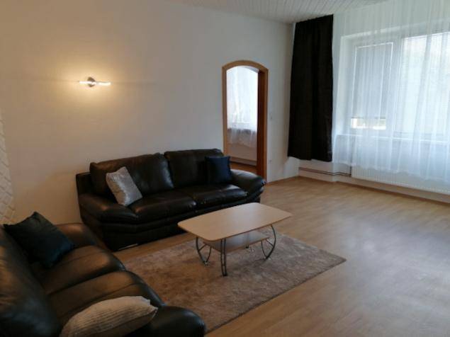 Gemütliches Wohnzimmer mit dunklen Sofas, hellem Laminatboden und großen Fenstern für viel Tageslicht.