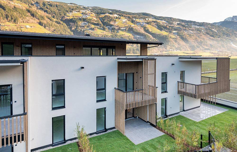 Ansicht einer modernen Wohnanlage mit Holzverkleidung, großzügigen Balkonen und Terrassen vor einer grünen Berglandschaft.