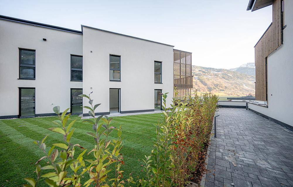 Gepflegte Rasenfläche mit Hecke und gepflastertem Weg vor der modernen Gebäudefassade mit großen Fenstern.