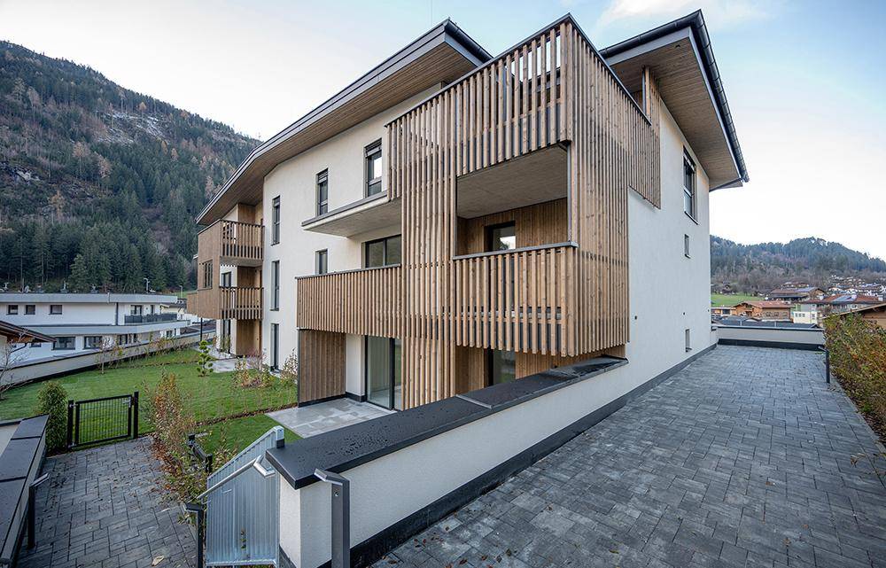 Eckansicht eines modernen Wohngebäudes mit Holzverkleidung, Balkonen und einem gepflasterten Zufahrtsbereich.