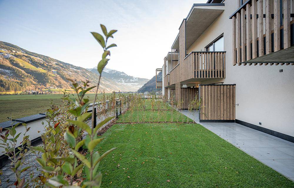 Gepflegte Grünflächen und Terrassen vor modernen Wohngebäuden mit Holzelementen und Bergblick.