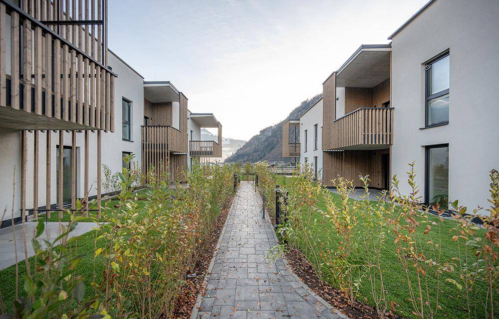 Moderne Wohngebäude mit Holzfassaden und Balkonen, getrennt durch einen gepflasterten Weg und grüne Hecken.