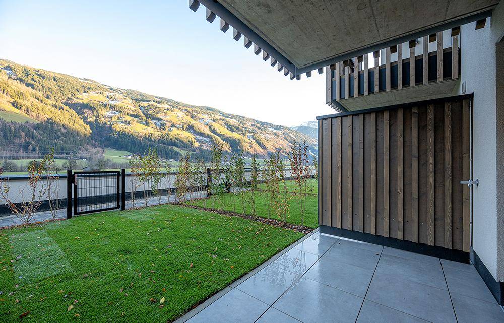 Sonniger Gartenbereich mit gepflasterter Terrasse und Rasenfläche, umgeben von einer Hecke und einem Zaun.