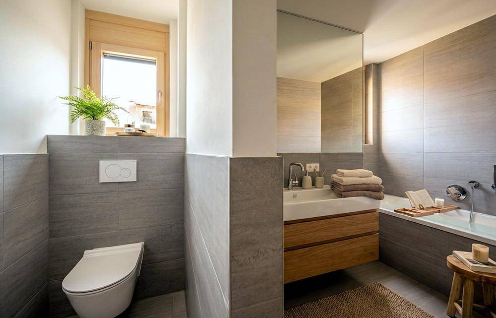 Modernes Badezimmer mit grauen Fliesen, Badewanne, wandmontierter Toilette und Fenster.
