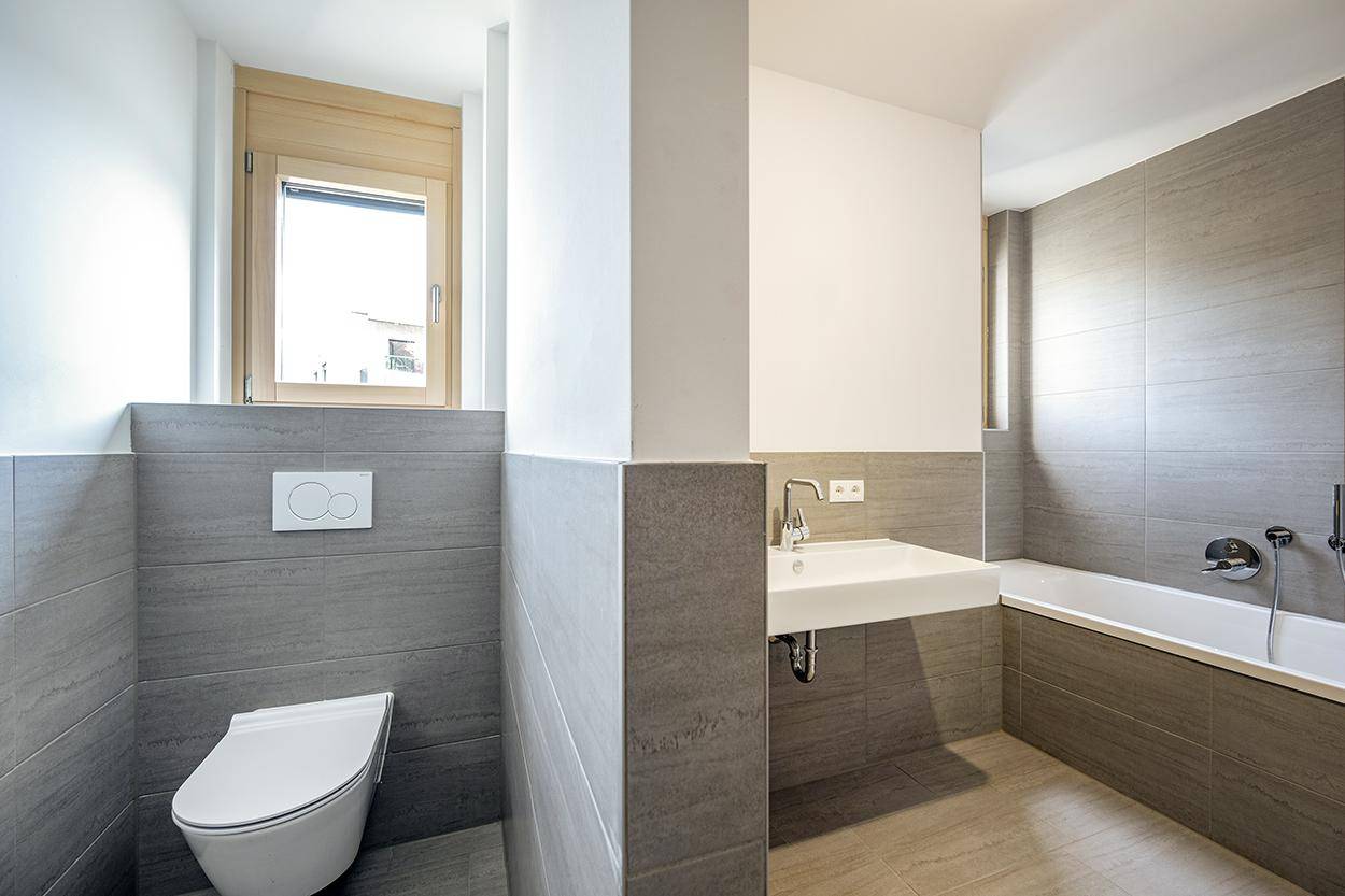 Helles Badezimmer mit Badewanne, wandmontierter Toilette, modernem Waschtisch und Fenster.