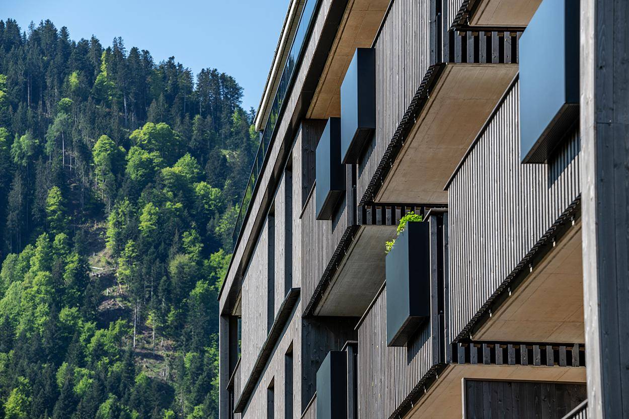 Detailansicht der modernen Holzfassade mit Balkonen und Fenstern vor einem bewaldeten Hang.
