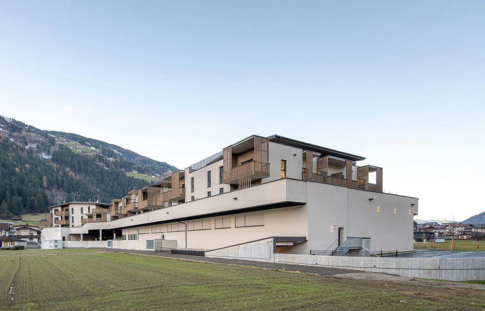 Moderne Wohnanlage in alpiner Landschaft mit weitläufigen Feldern im Vordergrund.