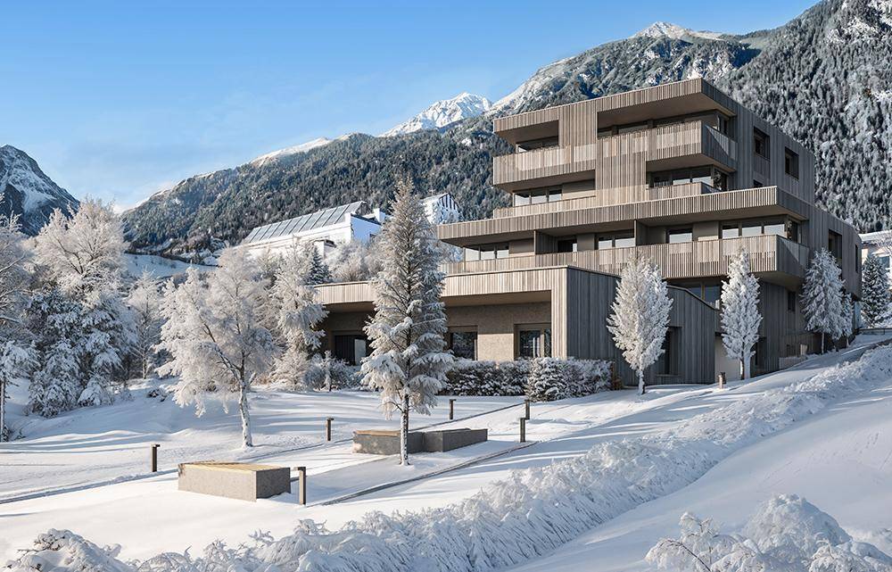 Moderne Wohnanlage in einer verschneiten Winterlandschaft mit Bäumen und Bergen im Hintergrund.