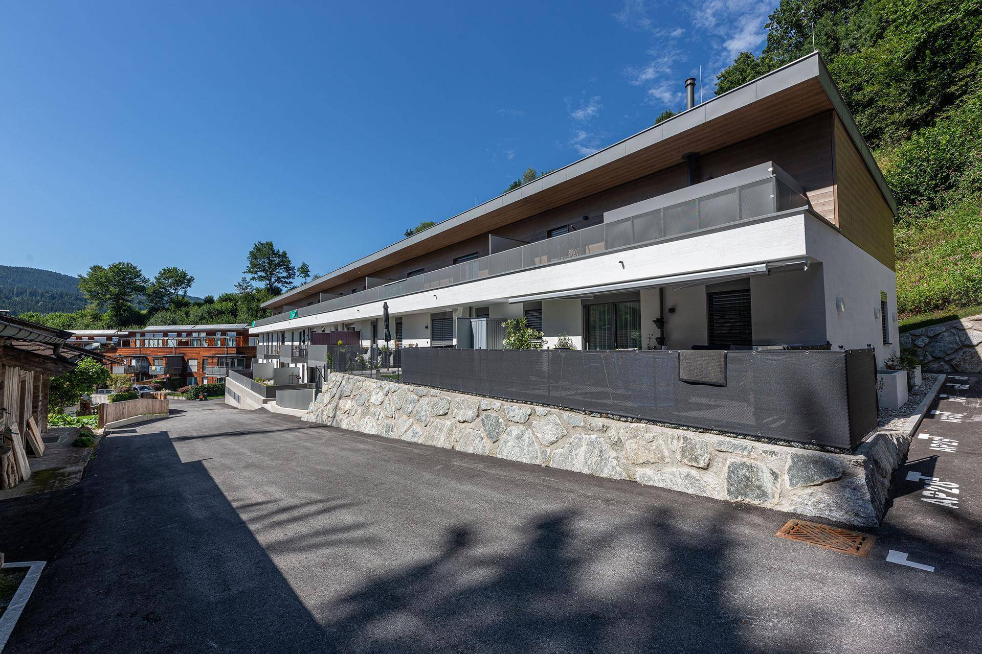 Modernes Mehrfamilienhaus mit Balkonen, umgeben von grüner Natur und Parkplätzen im Freien.
