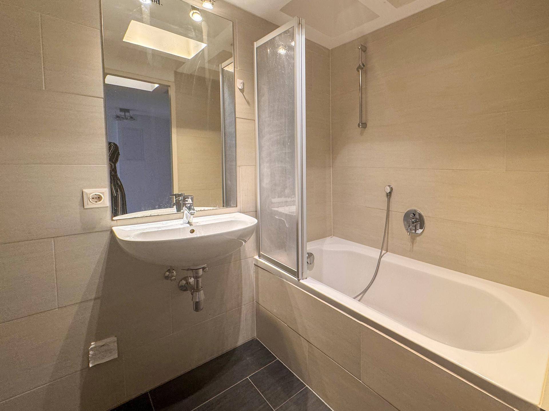 Helles Badezimmer mit Badewanne, Waschbecken und beigefarbenen Fliesen, ausgestattet mit Duschwand.