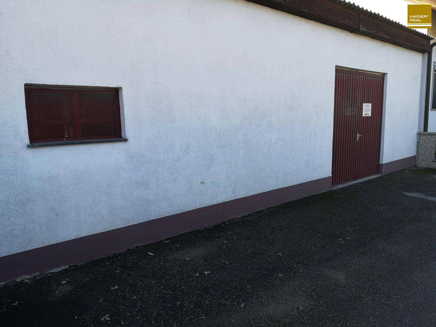 Außenansicht der Garage mit weißer Fassade, rotem Tor und vergittertem Fenster.