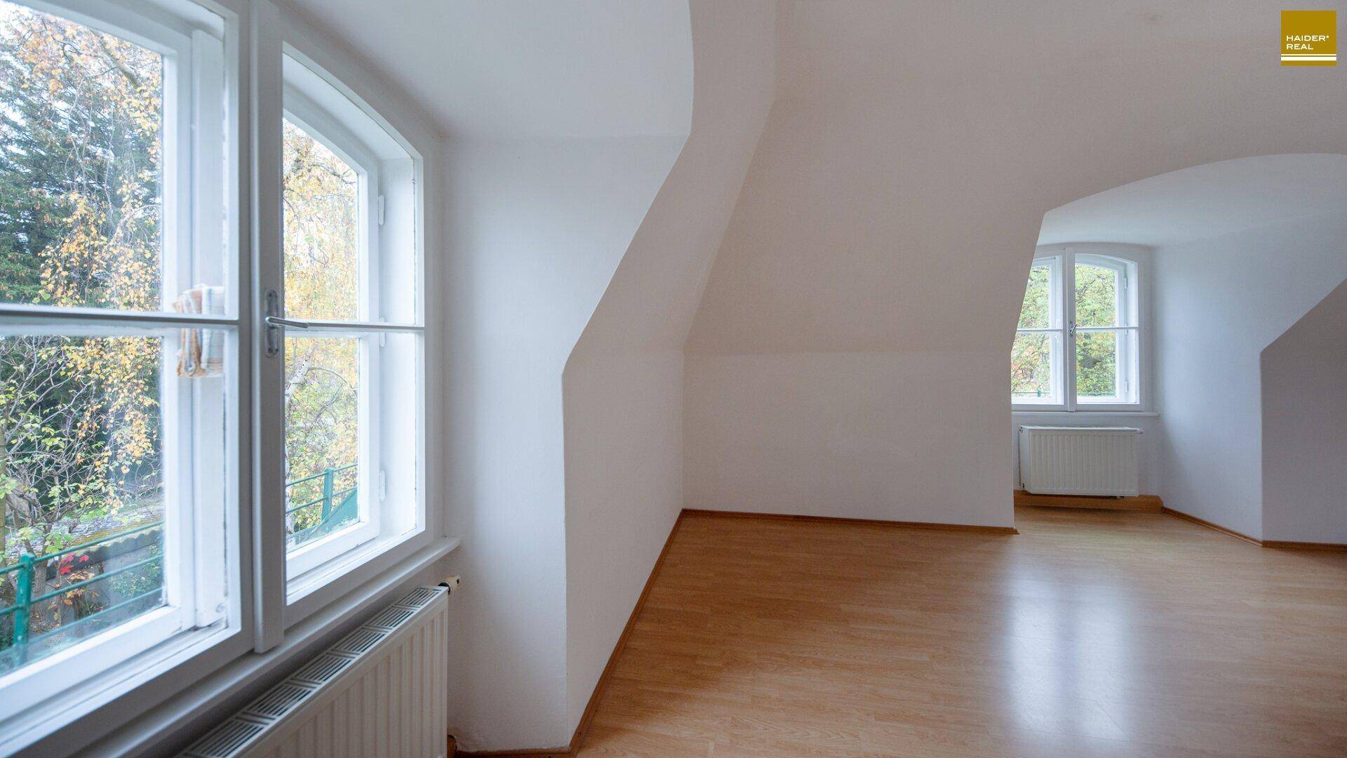 Helles Dachgeschosszimmer mit schrägen Wänden, Holzfußboden und mehreren Fenstern.