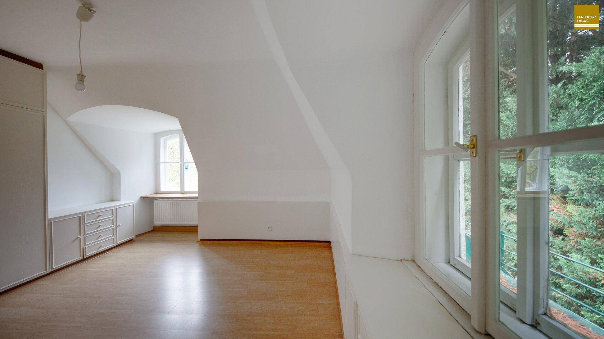 Geräumiges Dachgeschosszimmer mit schrägen Wänden, Einbauschränken und Blick ins Grüne.