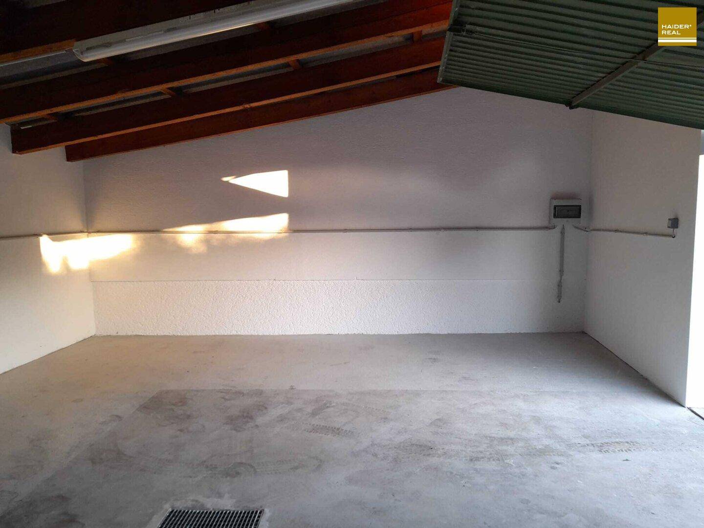 Helle Garage mit Betonboden, sichtbaren Holzbalken und elektrischen Installationen an der Wand.