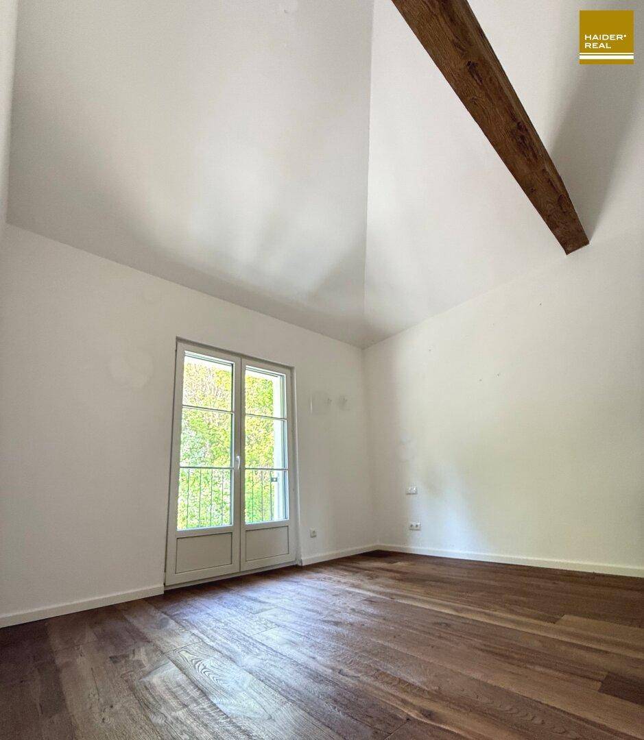 Helles Zimmer mit Holzboden, hohen Decken und einer Fenstertür, die viel Licht hereinlässt.