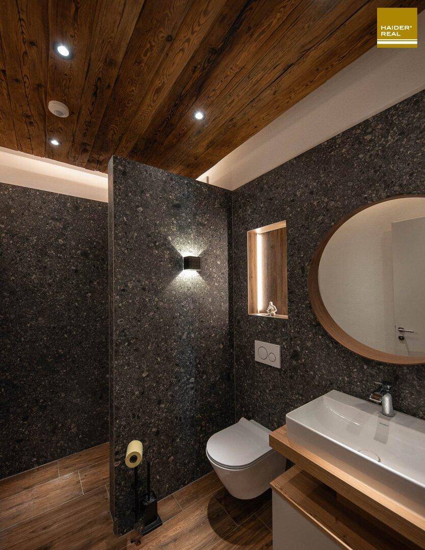 Modernes Gäste-WC mit dunklen Fliesen, Holzdecke und stilvollem Waschbecken mit rundem Spiegel.