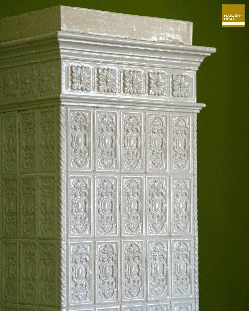 Weißer, reich verzierter Kachelofen mit traditionellen Mustern vor einer grünen Wand.